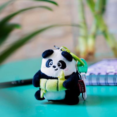 Panda-Plüsch-Schlüsselanhänger mit Bambus, aufrecht auf einem türkisfarbenen Tisch vor bunten Gegenständen dargestellt.