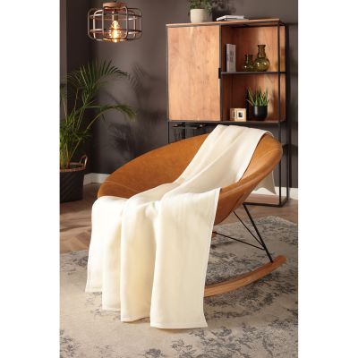 Wohndecke CottonSense natur fällt in langen cremefarbenen Falten über einen cognacfarbenen Loungesessel.
