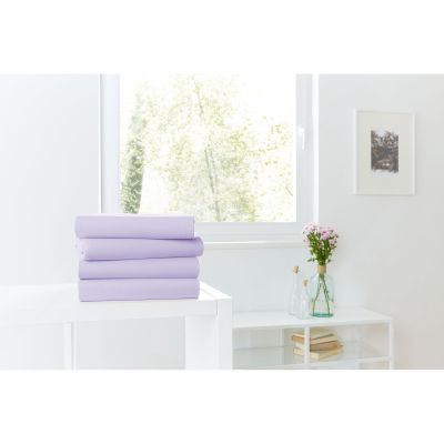 Vier gefaltete Spannbetttücher in Lavendel auf einer weißen Kommode vor einem hellen Fenster; Größe 100 x 200 x 30 cm.