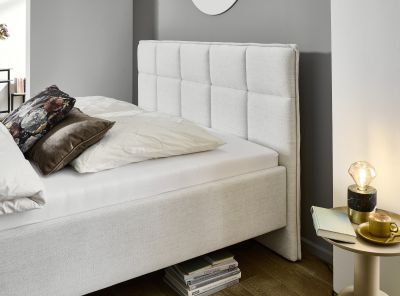 Detailaufnahme eines hellen Polsterbetts mit gestepptem Kopfteil, weißen Bettwaren und Kissen in einem modern eingerichteten Schlafzimmer