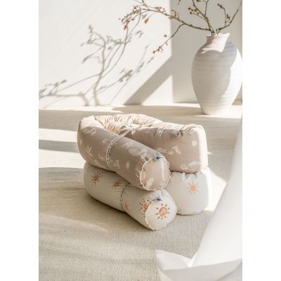 Zwei zusammengerollte Baby Bettschlangen in beige mit Tiermotiven und creme mit Sonnenmotiv, auf hellem Teppich neben einer weißen Vase im Sonnenlicht