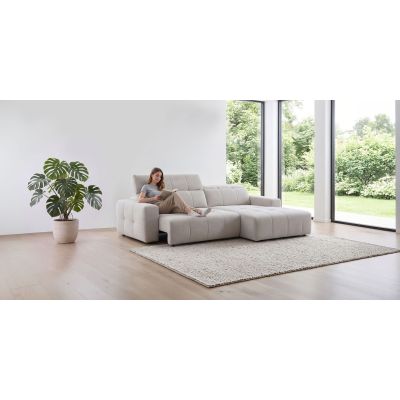 Person sitzt auf einer hellen Eckgarnitur mit Longchair rechts in einem Wohnraum mit Teppich, Pflanze und großen Fenstern