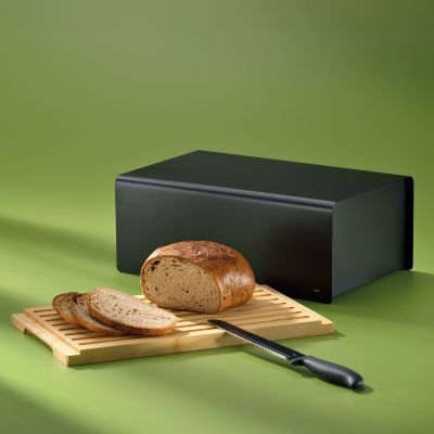 Schwarzer Brotkasten mit Brotlaib Messer und herausgezogenem Bambusbrett auf grünem Untergrund