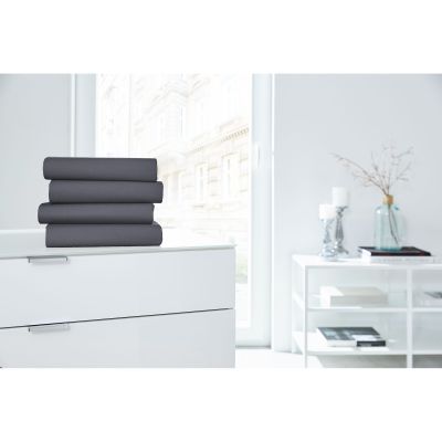 Vier gefaltete Boxspring-Spannbetttücher in Anthrazit auf einer weißen Kommode in einem hellen Raum; Größe 150 x 200 x 38 cm.