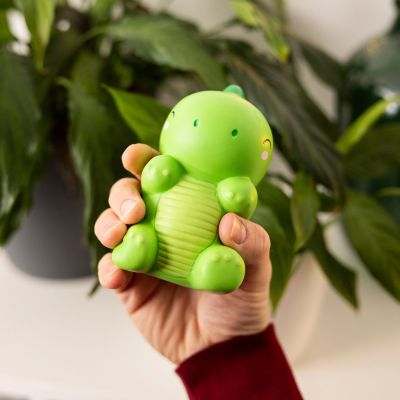 Grüne Antistress-Figur in Dinosaurierform, in einer Hand vor grünen Zimmerpflanzen gezeigt.