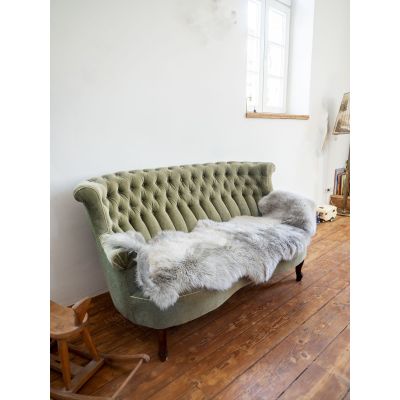 Großes graubeiges Lammfell liegt quer über Sitzfläche und Armlehne eines grünen, gesteppten Sofas in einem hellen Zimmer.