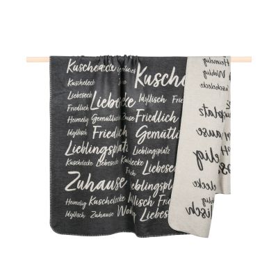 Graue Decke Snug, 150 × 200 cm, mit großflächigem schwarz-weißem Typografie-Muster, über einer Holzstange drapiert.