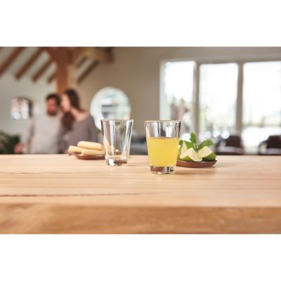 Zwei Schnapsgläser auf einem Holztisch eines mit gelbem Getränk und Limettenspalten daneben im Hintergrund Personen