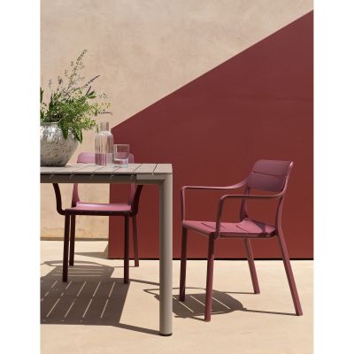 Zwei bordeauxrote Stühle mit Armlehnen stehen an einem rechteckigen Outdoor-Tisch. Auf dem Tisch stehen Gläser, eine Karaffe und eine Blumenvase, im Hintergrund eine beige-rote Wand.