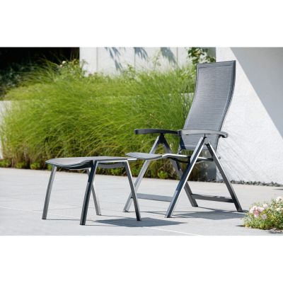 Ein grauer Gartenstuhl mit passendem Hocker steht auf einer sonnigen Terrasse neben einer weißen Hauswand. Im Hintergrund wachsen grüne Gräser und einige Blütenpflanzen.