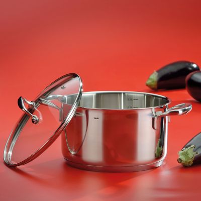 Geöffneter silberfarbener Bratentopf mit danebenliegendem Glasdeckel und Küchenutensilien auf rotem Hintergrund