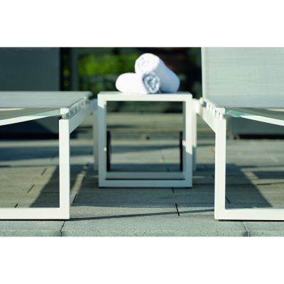 Ein weißer quadratischer Beistelltisch steht zwischen zwei Liegen auf einer Terrasse, auf dem Tisch liegen zwei zusammengerollte weiße Handtücher. Im Hintergrund ist unscharf Grün zu sehen.