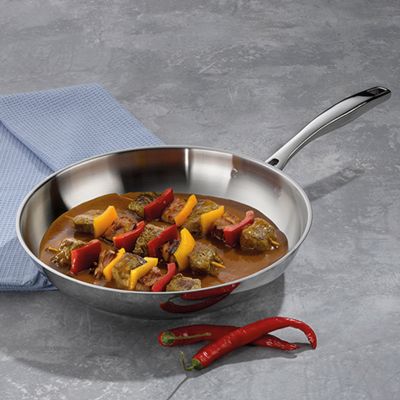 Bratpfanne aus Edelstahl mit gegartem Gemüse und Soße auf einer grauen Arbeitsfläche neben roten Chilischoten