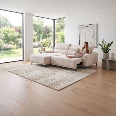 Beigefarbene Eckgarnitur Moon mit sitzender Person in einem hellen Wohnzimmer mit Teppich, Beistelltisch und Fensterfront