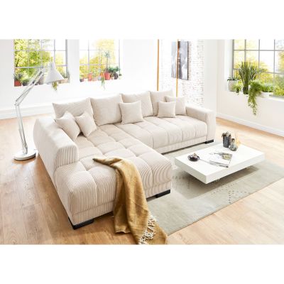 Cremige Wohnlandschaft aus Cord mit Longchair links im Bild, zahlreichen Kissen und beigefarbener Decke in einem hellen Wohnzimmer mit großen Fenstern