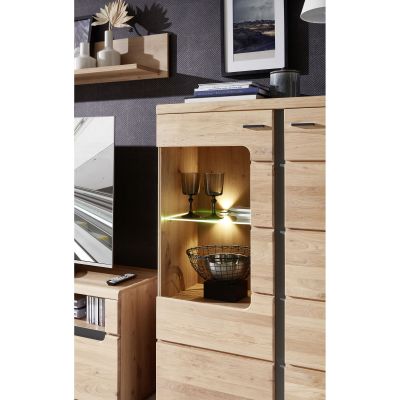 Detailaufnahme einer Vitrine in Holzoptik. Zu sehen sind zwei beleuchtete Glasböden mit Gläsern und einer dekorativen Metallschale. Im Hintergrund hängt ein Wandboard mit Vasen und einem Bild.