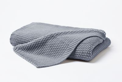 Gefaltete Strickdecke in Mittelgrau mit locker übergelegter Ecke; die grobe Waffelstrickstruktur ist vor hellem Hintergrund erkennbar; Größe 130 x 170 cm.