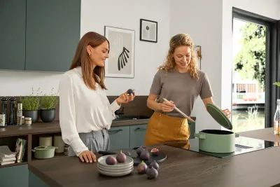 Zwei junge Frauen kochen in klassischer Interliving Küche mit graugrünen Fronten, dunklen Holzarbeitsflächen und Kücheninsel