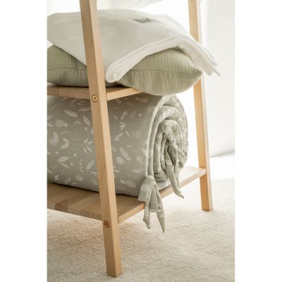 Zusammengerolltes olivgrünes Babybett-Nestchen mit weißem Blättermuster liegt auf einem Holzregal unter gefalteten weißen und grünen Textilien