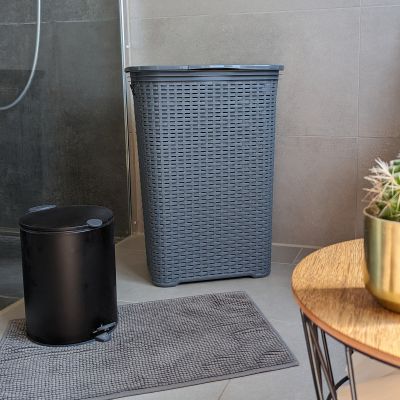 Rechteckige Wäschebox mit Flechtstruktur auf einem Badezimmerboden neben einem Abfalleimer, einem Teppich und einem Holzhocker