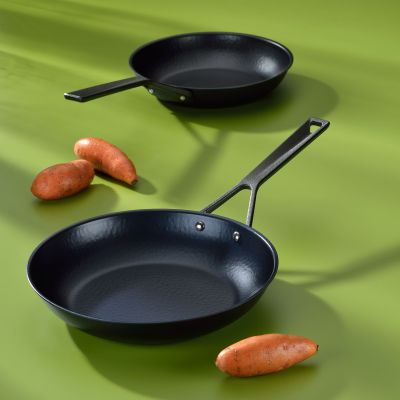 Zwei unterschiedlich große schwarze Bratpfannen mit langen Metallgriffen neben orangefarbenem Gemüse auf grüner Fläche