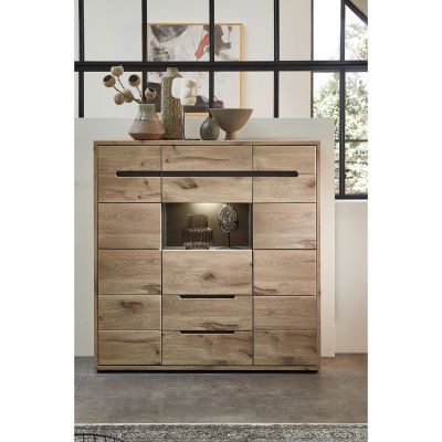 Die Kommode steht vor einer hohen schwarzen Fensterwand. Auf der Oberseite stehen verschiedene Dekoobjekte. Die Glasnische in der Mitte ist beleuchtet.