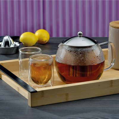 Gefüllte transparente Teekanne neben zwei Teegläsern und gelben Früchten auf einem Holztablett