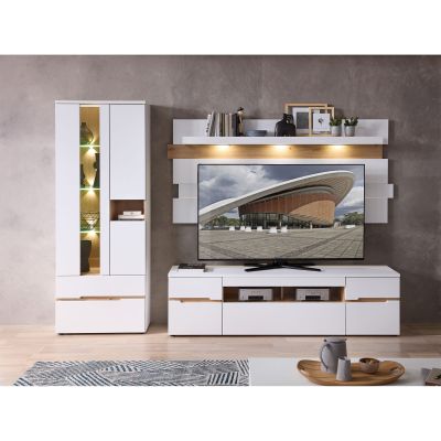 Wohnzimmeransicht mit einer weißen Wohnwand. Links steht eine Vitrine mit beleuchteten Glasfächern. In der Mitte befindet sich ein TV-Lowboard mit offenem und geschlossenem Stauraum. Dahinter sind weiße Wandpaneele mit einem Wandboard in Holzoptik und Bel