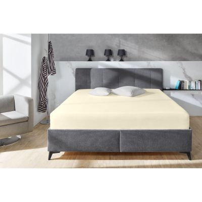 Boxspring-Spannbetttuch in Ecru auf einem grauen Boxspringbett in einem Schlafzimmer; zwei helle Kissen liegen am Kopfende; Größe 100 x 200 x 38 cm.