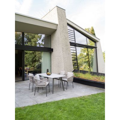 Eine moderne Terrasse mit vier beigen Stühlen aus Seilgeflecht und einem rechteckigen Tisch mit heller Platte. Die Terrasse liegt vor einem Haus mit großen Glasfronten und Blick auf grüne Bäume.