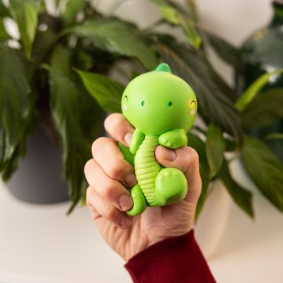 Grüne Antistress-Figur in Dinosaurierform, in einer zweiten Perspektive vor den Pflanzen gehalten gezeigt.
