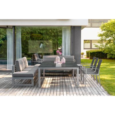 Eine Terrasse mit grauer Sitzgruppe bestehend aus zwei Bänken mit Rückenlehne, einem Tisch und mehreren Stühlen. Auf dem Tisch stehen Glasvasen mit Blumen, im Hintergrund sind Glasfronten und ein Garten mit Rasen und Bäumen zu sehen.