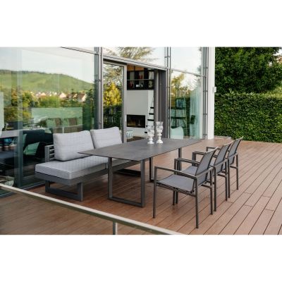 Eine Terrasse mit einer Sitzgruppe aus Metall und grauen Polstern. Eine Bank steht an einem Tisch mit vier passenden Stühlen. Im Hintergrund ist eine Glasfassade mit Blick ins Haus und auf grüne Hügel.