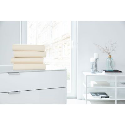 Vier gefaltete Boxspring-Spannbetttücher in Ecru auf einer weißen Kommode in einem hellen Raum; Größe 100 x 200 x 38 cm.