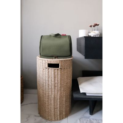 Kulturbeutel von Reisenthel, Modell Toiletbag XL, in Dunkelgrün mit goldfarbenen Metallteilen, geschlossen auf einem geflochtenen Korb in einem Wohnraum.