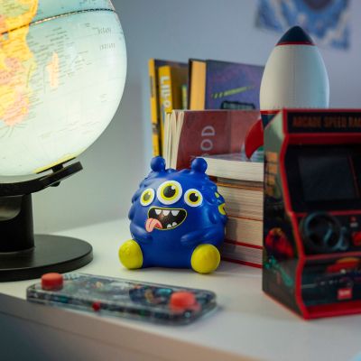 Blaue Antistress-Figur im Monster-Design, auf einem Schreibtisch neben Globus, Büchern und Mini-Arcade dargestellt.