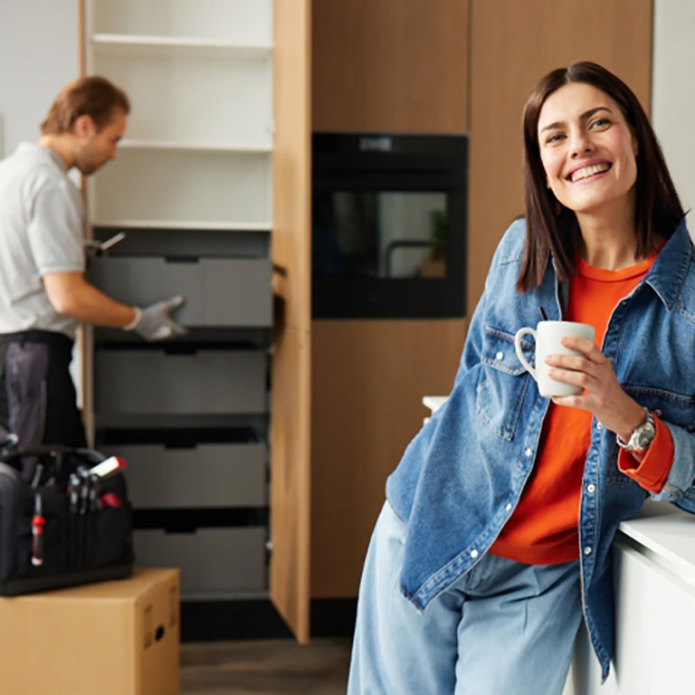 Lächelnde Frau mit einer Tasse, an die Küchentheke gelehnt; im Hintergrund arbeitet ein Mann an Schränken.