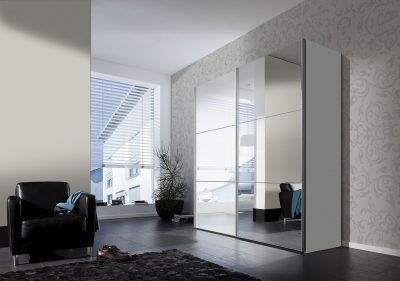 Modernes Zimmer mit Schwebetürenschrank von Möbel Gruber, schwarzem Sessel, Schuhen auf dem Boden und Fenster mit Jalousien.