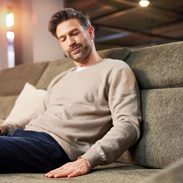 Mann, der einen beigen Pullover und dunkle Hosen trägt, sitzt gemütlich auf einem grünen Sofa in einem sanft beleuchteten Raum.