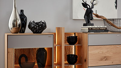 Die modernen Holzregale von Möbel Gruber präsentieren schwarze und silberne Vasen, Bücher und abstrakte Kunst an einer grauen Wand.