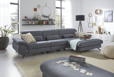 Modernes Wohnzimmer mit einer Polstergarnitur von Möbel Gruber, Kissen, Pflanzen, Wandkunst und großen Fenstern.