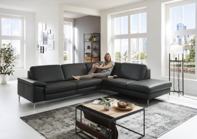 Eine Frau sitzt auf der Eckkombination, einem modernen schwarzen Sofa von Möbel Gruber, in einem hellen Wohnzimmer mit Stadtblick.