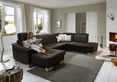 Frau entspannt auf einem großen Modulmaster Kiano-Sofa von Möbel Gruber in einem gemütlichen Raum mit Kamin und großen Fenstern.