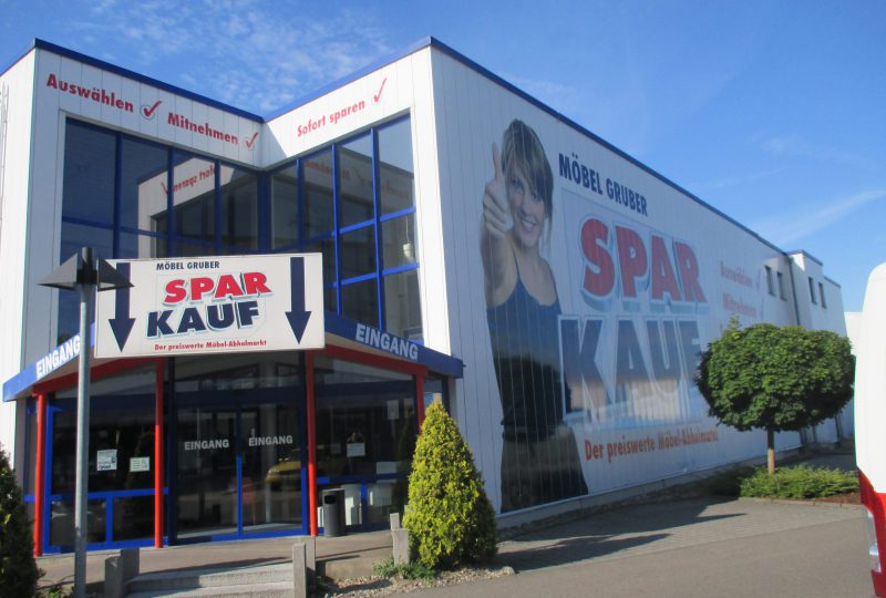 Die Außenansicht eines Möbel Gruber Spar Kauf-Geschäfts präsentiert sein großes Schild und seine Glasfassade unter einem klaren blauen Himmel.