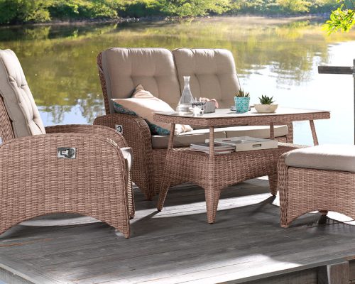 Das geflochtene Terrassenset von Möbel Gruber mit beigen Kissen auf einer Holzterrasse bietet Blick auf einen von üppigem Grün umgebenen Teich.