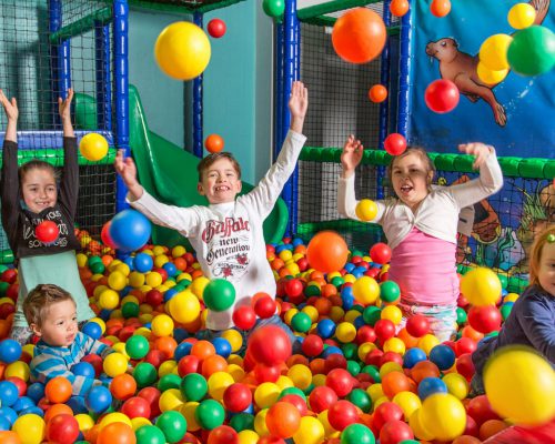 Fünf Kinder spielen fröhlich in einem Bällebad, umgeben von bunten Plastikbällen, einige Bälle schweben in der Luft und sie haben ein Lächeln im Gesicht.