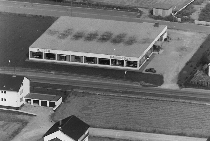 Luftaufnahme eines rechteckigen Gebäudes mit der Aufschrift „BRUMER“ mit umliegenden Straßen, Feldern und geparkten Autos. Möbel Gruber-Stil. Schwarzweißfoto.