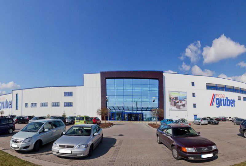 Großes Industriegebäude mit Glaseingang, „Möbel Gruber“ und parkenden Autos. Heller Tag, wenige Wolken.