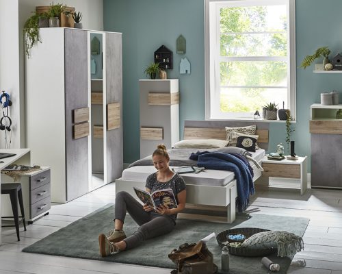 Eine Person sitzt auf einem Teppich und liest eine Zeitschrift in einem hellen Schlafzimmer mit modernen Möbeln und Dekor und einem großen Fenster.