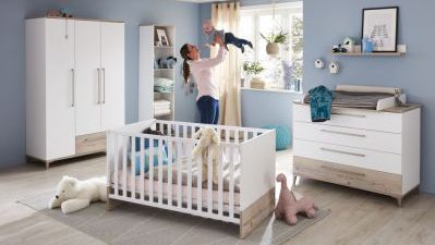 Eine Frau hält ein Baby in einem pastellfarbenen Kinderzimmer mit Kinderbett, Kommode, Stofftieren und Kleiderschrank.